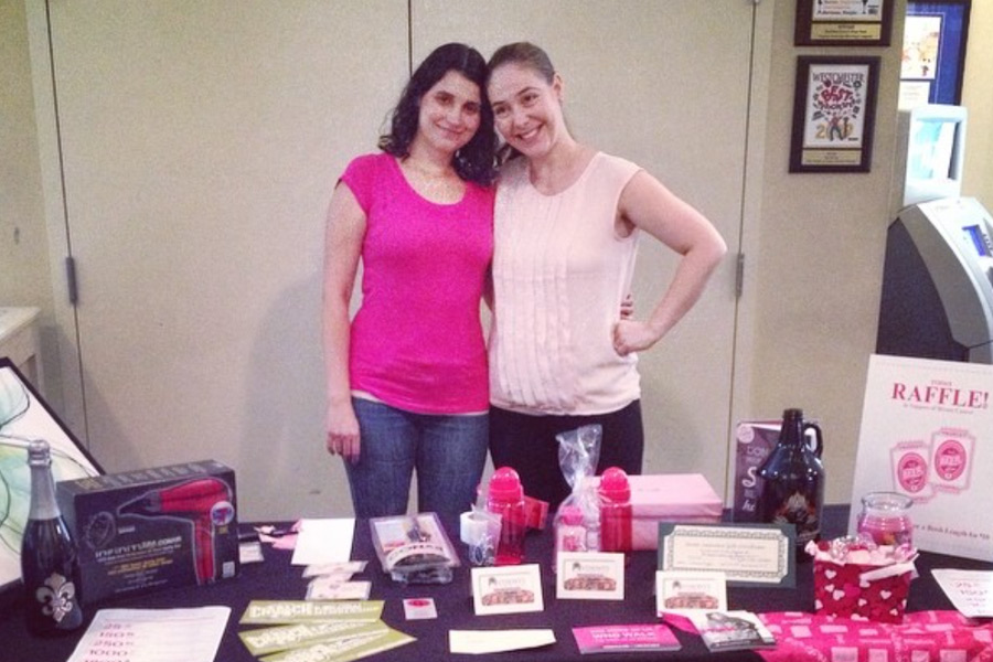 Two women in pink smiling behind a Raffle table